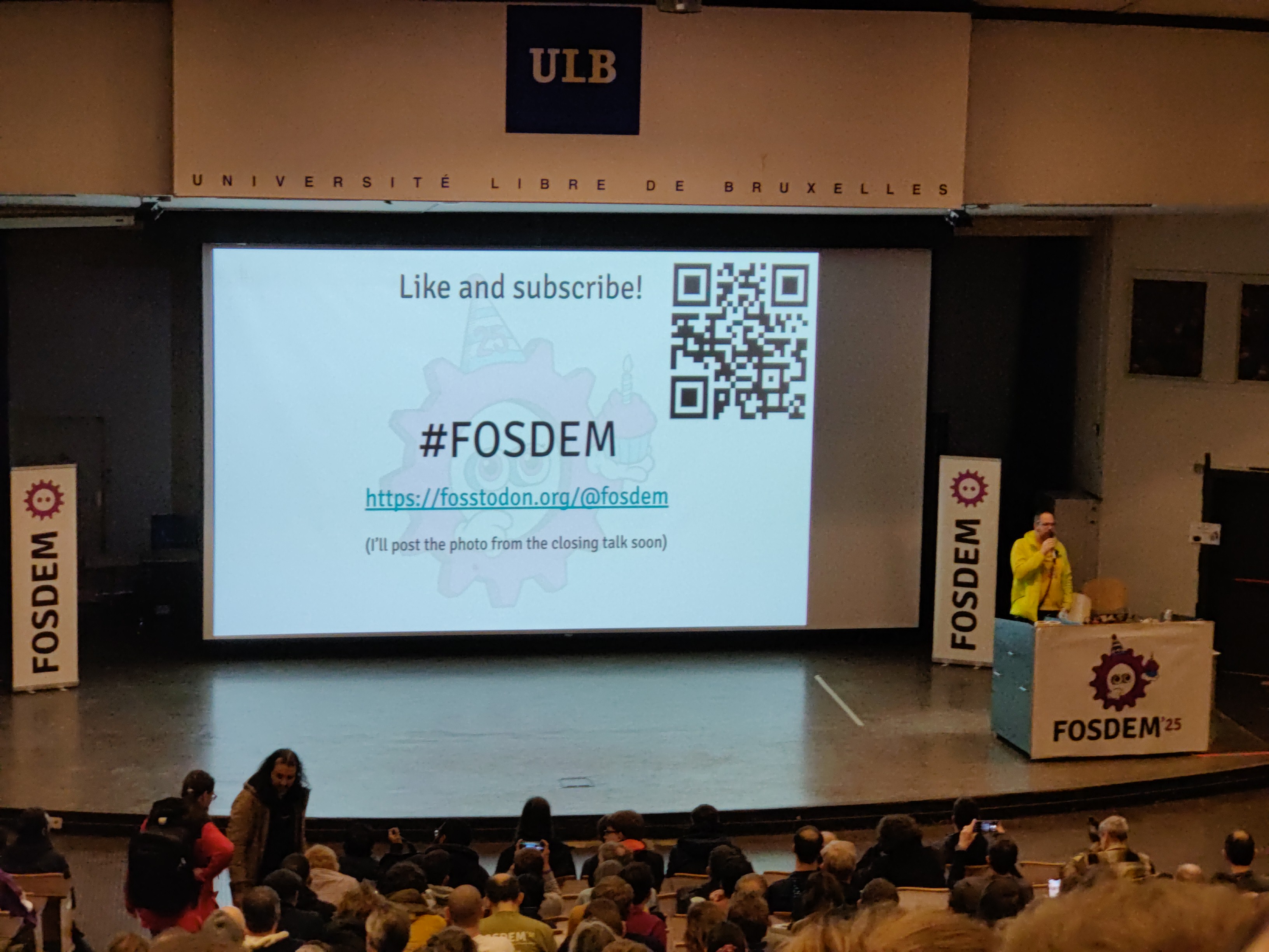 Foto al talk conclusivo del FOSDEM 2025. Si vede ripetuto il logo del FOSDEM (un ingranaggio viola con 2 punti nel mezzo) Foto al talk conclusivo del FOSDEM 2025. Si vede ripetuto il logo del FOSDEM (un ingranaggio viola con 2 punti nel mezzo)